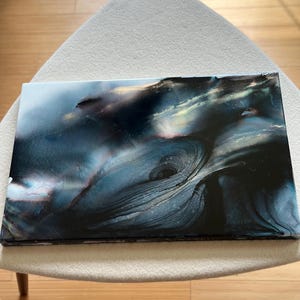 May include: Abstract art piece with a dark, swirling design in shades of blue, black, and white. The artwork has a glossy finish and is rectangular in shape. The piece is displayed on a light-colored chair.