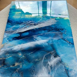 May include: Abstract ocean-themed artwork with shades of blue, white, and gold. The painting features a glossy finish, with fluid patterns resembling waves and sea foam. The artwork is rectangular and appears to be mounted on a wooden frame.