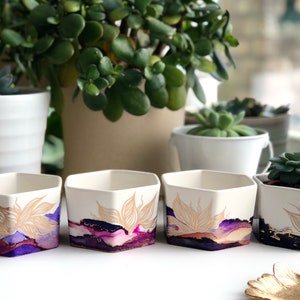 May include: Four hexagonal ceramic planters featuring a white base with abstract purple, magenta, and dark blue ink-style washes. Each planter is decorated with delicate, gold-colored line art of a stylized flower petal design on the front.