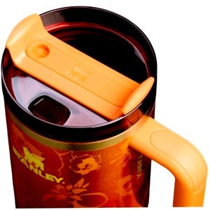 May include: A Stanley brand travel mug with a vibrant orange and red floral pattern. The mug features a dark brown lid with a bright orange sliding closure mechanism and a matching orange handle extending from the side of the cylindrical body.