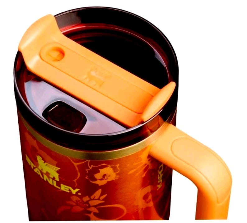 May include: A Stanley brand travel mug with a vibrant orange and red floral pattern. The mug features a dark brown lid with a bright orange sliding closure mechanism and a matching orange handle extending from the side of the cylindrical body.