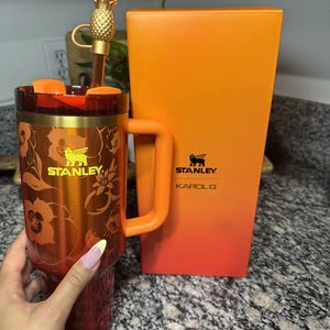 May include: A red and orange Stanley tumbler with a floral pattern, a gold-colored Stanley logo, and a matching orange handle. The tumbler includes a straw topped with a gold pineapple charm and is displayed next to its orange and red gradient box.
