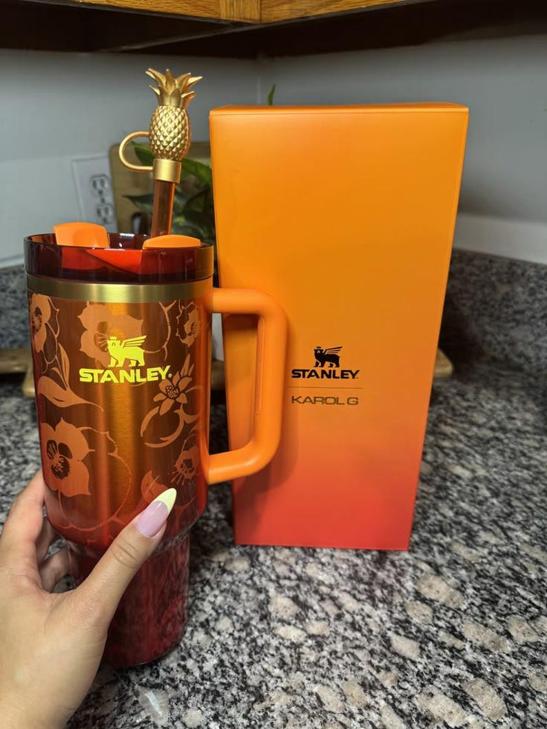 May include: A red and orange Stanley tumbler with a floral pattern, a gold-colored Stanley logo, and a matching orange handle. The tumbler includes a straw topped with a gold pineapple charm and is displayed next to its orange and red gradient box.