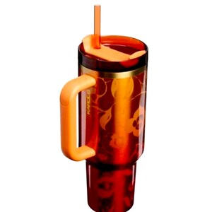 May include: A translucent deep red tumbler featuring a floral pattern, a solid orange handle, a matching orange lid, and a clear orange straw. The side of the tumbler is embossed with the text KAROL G in a vertical orientation near the handle.