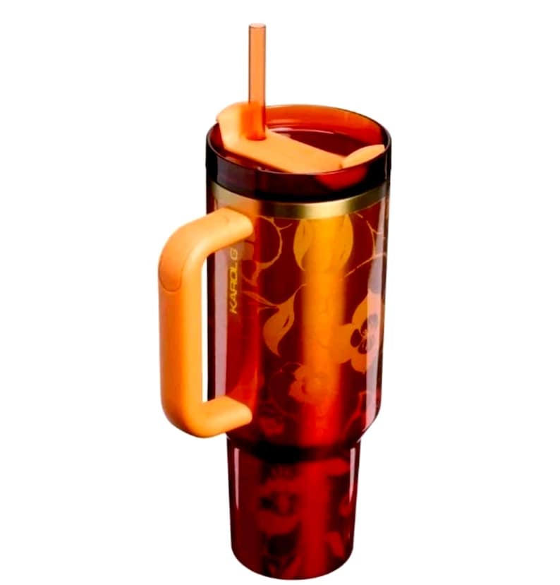 May include: A translucent deep red tumbler featuring a floral pattern, a solid orange handle, a matching orange lid, and a clear orange straw. The side of the tumbler is embossed with the text KAROL G in a vertical orientation near the handle.