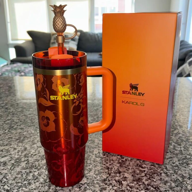 May include: A metallic orange and red Stanley tumbler with a floral pattern, a side handle, and a straw topped with a gold pineapple charm. The tumbler features the Stanley logo and is accompanied by a matching orange and red rectangular box.