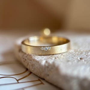 May include: A gold band ring with a brushed matte finish featuring a small, semi-circular cutout detail on the top surface. This cutout is set with three tiny, clear, round-cut stones arranged in a horizontal row along the band's edge.