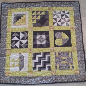 Op de afbeelding: Een handgemaakte patchworkquilt met een geometrisch ontwerp. De quilt heeft negen vierkanten met patronen in grijs, zwart en wit, tegen een lichtgroene achtergrond. De rand is een gespikkelde grijze stof. De quilt is ongeveer 100 cm vierkant.