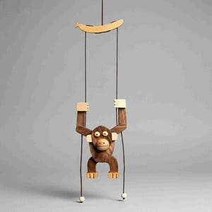 May include: A wooden monkey toy suspended from a string, featuring a banana-shaped element at the top. The monkey is brown with light-coloured accents on its hands and feet. The toy hangs against a grey background.