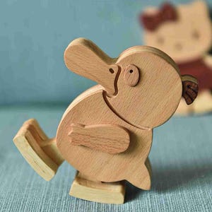 May include: A light-coloured wooden figurine shaped like a platypus with a flat bill, a rounded body, and small flipper-like appendages. The figure features a visible wood grain texture and is constructed from several interlocking wooden pieces.