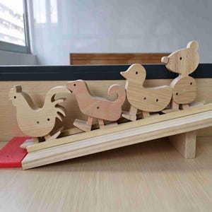 Könnte beinhalten: Ein Set aus vier hellen Holzfiguren in Form eines Hahns, eines Hundes, einer Ente und einer Maus, die in einer Reihe auf einer Holzrampe stehen. Jede Figur hat ein einfaches Ausschnitt-Design mit einem kleinen runden Auge und glatter Oberfläche.