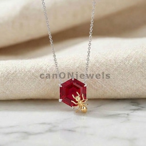 Peut inclure: Un collier en argent avec un pendentif en forme d'hexagone rouge. Un charm en forme de fourmi dorée est attaché au pendentif. Le texte "canoNjewels" est visible en arrière-plan.