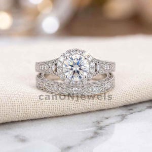 Peut inclure: Un ensemble de bagues de fiançailles en diamant avec une pierre centrale ronde et un halo de petits diamants. La bague présente des détails complexes et est associée à une alliance assortie. Le texte "canonjewels" est visible.