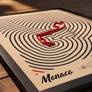 Può includere: Opera d'arte incorniciata con un contorno rosso di pista da corsa su linee nere concentriche. La parola "Monaco" è stampata in nero sotto. Una piccola illustrazione di un'auto si trova nell'angolo in basso a destra.