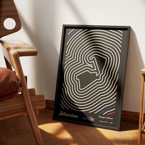 May include: Black framed art print featuring a white and black abstract design. The print includes the text "Silverstone" and "Inaugural Winner George Form - 1950". The artwork is leaning against a wall next to a wooden chair.