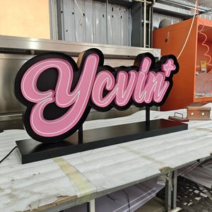 Könnte beinhalten: Ein individuelles 3D-Schild mit dem Wort Yevin+ in einer kursiven Schriftart. Die Buchstaben haben eine leuchtend rosa Vorderseite mit weißer Umrandung und sind auf einem schwarzen Hintergrund montiert. Das Schild steht auf einem schwarzen Sockel.