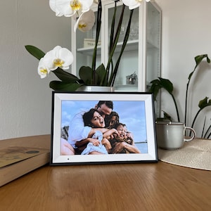 May include: A rectangular digital photo frame with a black border and a white inner bezel displaying a color photograph of a person embracing two children outdoors. The device features a small circular sensor on the bottom edge of the frame.