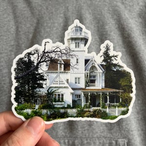 May include: A die-cut sticker featuring a white Victorian-style house with a tall tower, multiple gables, and a wrap-around porch. The house is surrounded by dark green trees and a white picket fence, with a bare tree branch extending over the roof.