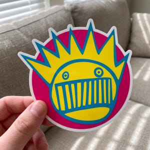 May include: A circular sticker featuring a yellow face with a wide, blue-outlined mouth showing vertical teeth, two small circular eyes, and a large, spiky yellow crown with blue outlines, all set against a solid magenta circular background.
