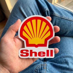 May include: A die-cut sticker featuring the Shell logo, which consists of a yellow scallop shell shape with a red outline. Below the shell graphic, the word Shell is printed in bold, red, sans-serif capital letters on a white background.