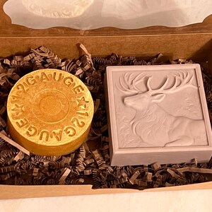 May include: A gift box containing two items: a round, golden-colored soap with "12 GAUGE" text and a rectangular, light-brown soap with a deer relief. The box is filled with brown paper shreds.