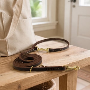 Puede incluir: Una correa de cuero marrón oscuro para perro con herrajes de latón, que incluye dos mosquetones giratorios y dos anillos de metal. La correa está enrollada junto a un bolso de lona beige y muestra costuras visibles a lo largo de la tira.