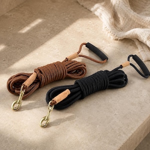 Peut inclure: Deux laisses pour chien enroulées, une marron et une noire, chacune dotée d'un design en corde tressée, d'une poignée rembourrée noire, d'un accent en cuir beige près de la poignée et d'un mousqueton pivotant en métal doré à l'extrémité.