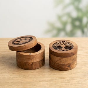 Op de afbeelding: Twee ronde houten bakjes met deksels. Eén deksel heeft een uitgesneden pootafdruk, terwijl het andere deksel een gedetailleerde gravure van een levensboom toont. De bakjes zijn gemaakt van natuurlijk bruin hout met zichtbare nerven.