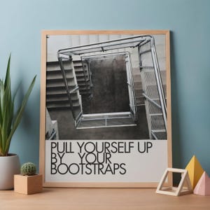 May include: Framed black and white print featuring a descending staircase with metal railings. The print includes the text "PULL YOURSELF UP BY YOUR BOOTSTRAPS" in a bold, sans-serif font. The frame is made of light-colored wood.