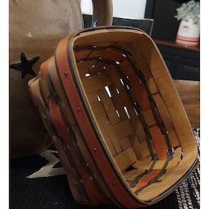 May include: A square woven wood basket featuring light tan, dark brown, and muted orange splints. The basket has a reinforced rim with visible metal tacks securing the overlapping strips of wood in a classic checkerboard weave pattern.
