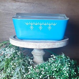 May include: A rectangular turquoise glass casserole dish with a clear glass lid. The exterior of the dish features a white printed pattern consisting of a scalloped line along the top edge with hanging starburst motifs spaced evenly across the side.