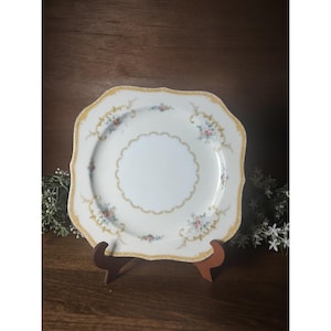 May include: A square porcelain plate with scalloped edges, featuring a white center, a gold filigree border, and delicate floral patterns in pink, blue, and green. The plate is displayed on a brown stand, showcasing its ornate design and fine details.
