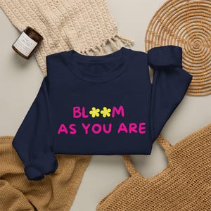 Könnte beinhalten: Ein marineblaues Langarm-Sweatshirt mit dem pinkfarbenen Schriftzug "BLOOM AS YOU ARE" auf der Vorderseite. Der Buchstabe "O" im Wort "BLOOM" ist durch zwei kleine, gelbe, fünfblättrige Blumen ersetzt. Das Sweatshirt hat einen Rundhalsausschnitt.