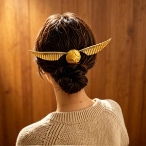 Puede incluir: Un accesorio para el cabello de color dorado con forma de esfera y dos alas emplumadas extendidas a los lados. El accesorio se coloca horizontalmente en la parte posterior del cabello, sujetando un moño en su lugar con su acabado metálico.