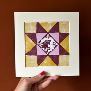 Könnte beinhalten: Ein quadratischer Blockdruck mit einem geometrischen Sternmuster in Senfgelb und Dunkelkastanienbraun. Das mittlere Rautenfeld zeigt eine stilisierte Pferdesilhouette in Kastanienbraun auf hellrosa Grund, umrahmt von einem weißen Passepartout.