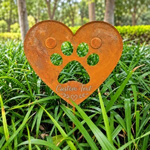 May include: A heart-shaped garden stake made of rusted metal featuring a cutout paw print design in the center. The surface includes the engraved text Custom Text above a small heart icon flanked by decorative leaf-like flourishes.