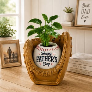 Könnte beinhalten: Ein Baseball-Themen-Pflanzgefäß mit einer grünen Pflanze im Inneren, eingebettet in einen braunen Baseballhandschuh. Der weiße Pflanztopf ist wie ein Baseball gestaltet und trägt den Text "Happy Father's Day" in Schwarz. Ein gerahmtes Foto und ein Schild mit der Aufschrift "Best Dad Ever" befinden sich im Hintergrund.