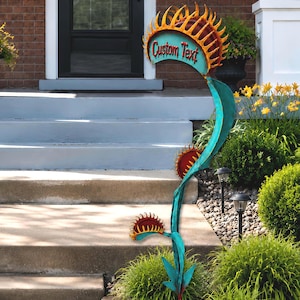 May include: A metal garden sculpture shaped like a Venus flytrap plant with a tall, teal-colored stem and three open traps. The traps feature jagged, orange-yellow edges and deep red interiors. The top trap displays the words Custom Text.