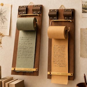 Könnte beinhalten: Zwei wandmontierte Papierabroller mit rechteckigen Brettern aus dunklem Holz, braunen Lederriemen und goldfarbenen Metallbeschlägen. Jede Einheit hält eine Papierrolle mit handschriftlichem Text und eine goldene Leiste mit der Aufschrift THOUGHTS IDEAS NOTES und WRITE THINK REMEMBER.