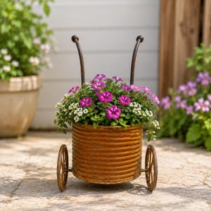 May include: A cylindrical, corrugated metal planter with a rust-colored finish, mounted on two small metal wheels with two curved handles extending upward. The planter is filled with vibrant purple petunias and small white alyssum flowers.