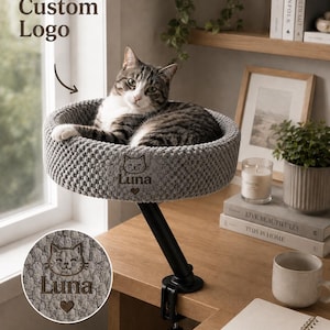 May include: A gray cat bed with the name "Luna" and a cat face design is mounted on a black adjustable arm. The cat bed is round and has a textured surface. A gray and white tabby cat is resting in the bed. The words "Custom Logo" are in the upper left corner.