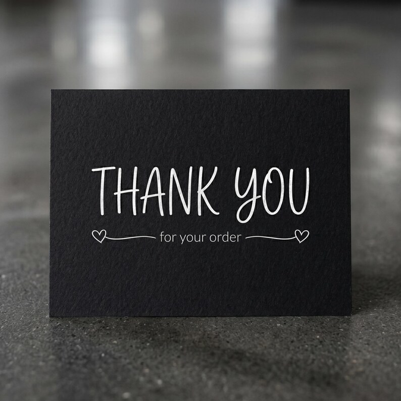 May include: A black card with the words "THANK YOU" in white script. Below the text is "for your order" in a smaller font. Two small heart shapes are on either side of the text.