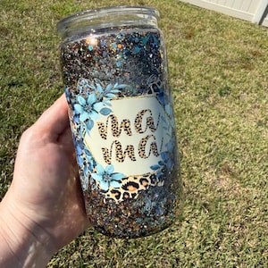 May include: A clear glass tumbler with glitter and floral patterns. The tumbler has the word "MaMa" in a leopard print font, framed by blue floral designs. The glitter inside is a mix of blue, silver, and brown. The tumbler is a stylish drinkware item.