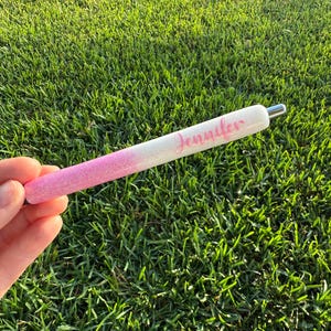 Puede incluir: Bol&iacute;grafo brillante con un dise&ntilde;o degradado de rosa a blanco. El nombre "Jennifer" est&aacute; escrito en cursiva en el bol&iacute;grafo. El bol&iacute;grafo tiene una punta de metal plateado. El fondo es hierba verde.