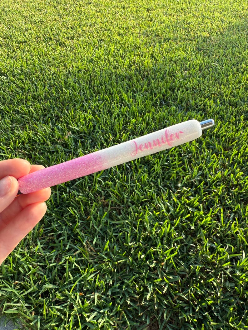 Puede incluir: Bol&iacute;grafo brillante con un dise&ntilde;o degradado de rosa a blanco. El nombre "Jennifer" est&aacute; escrito en cursiva en el bol&iacute;grafo. El bol&iacute;grafo tiene una punta de metal plateado. El fondo es hierba verde.
