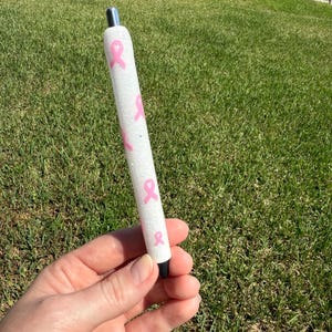 May include: A white pen with pink ribbon designs. The pen has a silver tip and a black tip. The pen is decorated with pink ribbons and glitter.