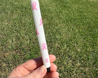 Può includere: Una penna bianca con disegni a nastro rosa. La penna ha una punta argentata e una punta nera. La penna è decorata con nastri rosa e glitter.