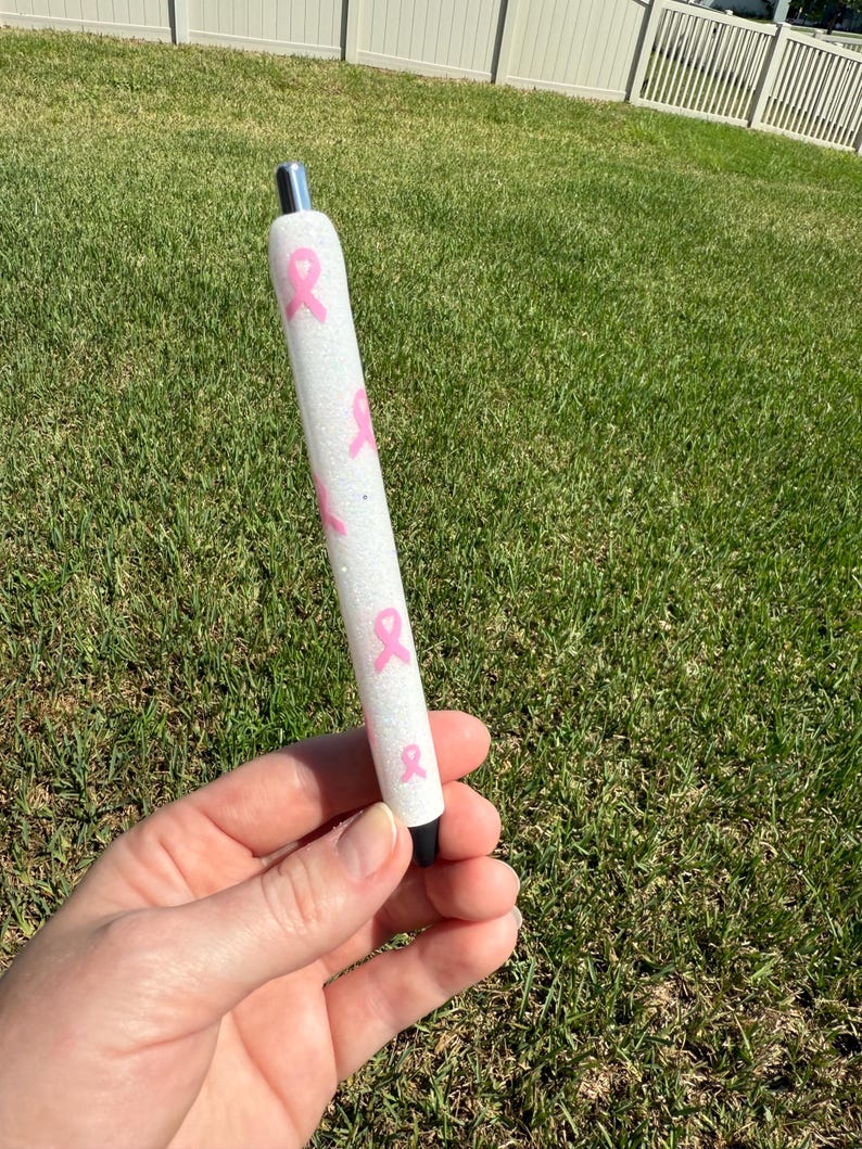 May include: A white pen with pink ribbon designs. The pen has a silver tip and a black tip. The pen is decorated with pink ribbons and glitter.