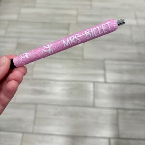 Puede incluir: Un bol&iacute;grafo rosa brillante con el texto "MRS. BILLET" en blanco. El bol&iacute;grafo tiene una punta plateada y una base negra. El bol&iacute;grafo se sostiene en una mano.