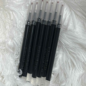 May include: A collection of black pen refills with clear plastic caps. The refills are arranged diagonally against a white, fluffy background. Each refill has a white base and a red tip.